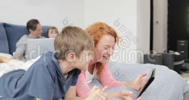 妈妈和儿子一起笑看数码平板电脑上的搞笑视频躺在床上，快乐的一家人在一起